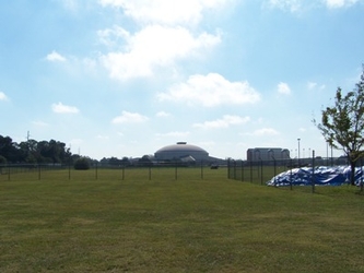 A large empty grass field bordered by chain-link fencing; trees and buildings are in the far background.