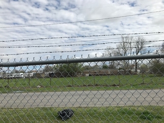 A chain-link fence with a barbed wire top in the foreground, a road running horizontally behind it, and grass on each side of the road, with another fence and trees on the far side.