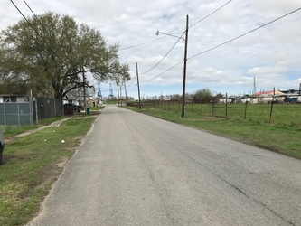 A road in the center extending toward the horizon, flanked by grassy areas with fencing, trees, and electrical poles around under a clouded sky.