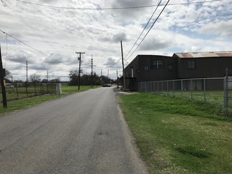 A road extending vertically into the distance with grass and fencing on either side; a large building is on the right, and trees and electrical poles are in the distance.