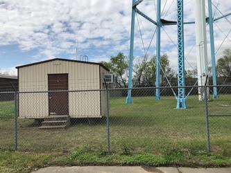 A small tan building to the left, a chain-link fence in the foreground, and the base of a water tower on the right, all in a grassy field with trees in the background under a cloudy sky.