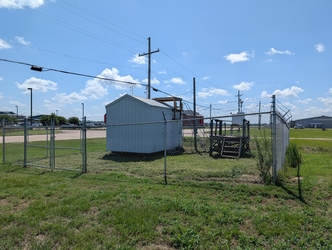A small white shed and a wooden platform with air monitoring equipment, both located inside a square of chain-link fencing on a grass field; roads, buildings, cars, and trees are in the background.