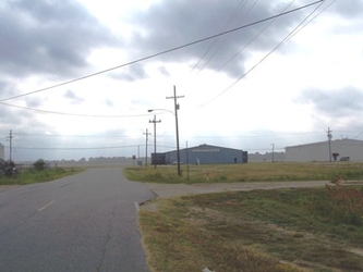 A road on the left extending toward the horizon flanked by fields on either side with a side road branching off to the right and buildings in the distance.