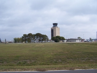 An expansive field with a tall structure in the far background; trees, cars, and buildings are in the far center under a cloudy sky.