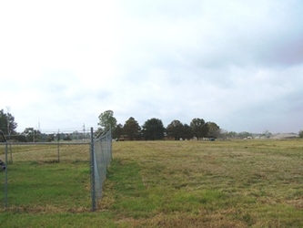 A vast grass field with part of a chain-link fenced-in area visible to the left and trees in the background under a clouded sky.