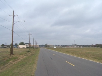 A road extending vertically from the foreground into the horizon, flanked by grass fields on either side, with electrical poles along the left side and trees in the background under a cloudy sky.