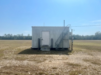 A small shed in the middle of a vast, barren field under a clear blue sky with a forest line on the horizon.