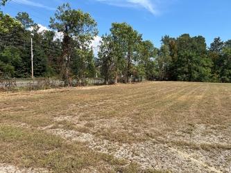 A wide open, barren field with trees all along the background and a chain-link fence on the left bordering a road on the outer side.