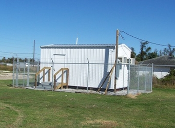 A small white building inside a chain-link-fenced plot of gravel, surrounded by short grass with trees in the background and a clear blue sky overhead.
