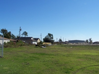 A vast grass field with various buildings, electrical lines, and trees in the background under a clear blue sky.