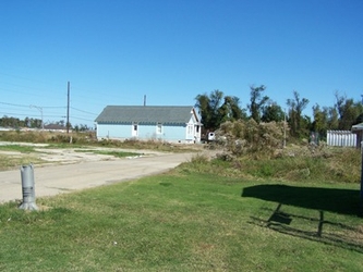 A grass field with pavement on the left, the shadow of a structure on the right, and a building and trees in the background under a clear blue sky.
