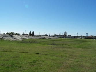 A vast grass field with a large mound of sediment on the distant left and trees lining the horizon under a clear blue sky.