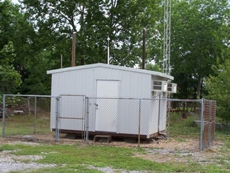 A small white building inside a chain-link-fenced area of grass and dirt with dense trees filling the background and view of the sky.