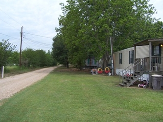 A grass yard in the foreground and center, a road extending vertically on the left, and a mobile home on the right, with trees filling the background under an overcast sky.