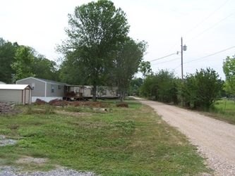 A grass yard on the left with a road extending vertically ahead on the right, a mobile home on the distant left, and trees throughout the background.