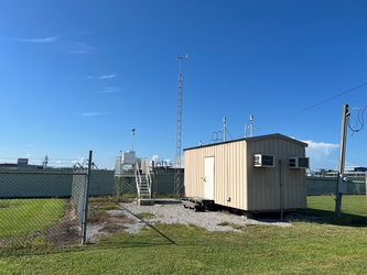 A small tan building and metal platform with air monitoring equipment to the right on a plot of gravel within a grass field; there is a solid barrier in the background.
