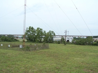 A vast grassy field with a fenced-off metal tower to the left and trees in the distance blocking a bridge under the cloudy sky.