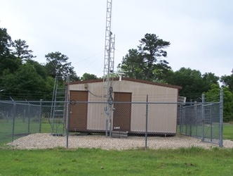 A small tan building inside a fenced-in gravel plot within a grass field with trees in the background under a cloudy sky.