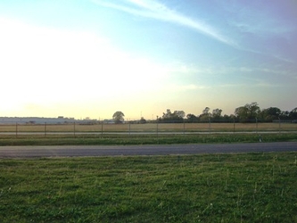 A road running horizontally down the middle with grass on either side, fencing on the far side, and trees in the distance under a colorful sky.