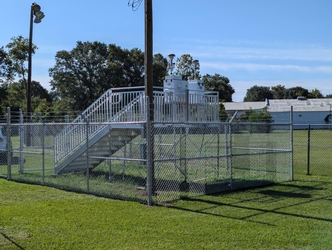 A metal platform with two air sampling instruments and a set of stairs inside a chain-link fenced-in area within a grass field; trees, electrical poles, and buildings are in the background.