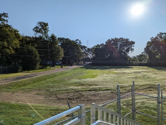 An empty grass field with part of metal railing and fencing in the foreground, a road on the left, and trees in the background under a clear, sunny sky.
