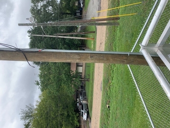 Part of a metal railing and chain-link fencing is in the foreground with an electrical pole close in the center, grass extends beyond the fencing to a dirt road that meets a residential property.