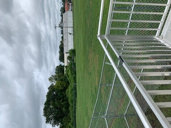 A lush grass field with part of a metal railing and chain-link fencing in the foreground and trees and buildings in the background under a clouded sky.
