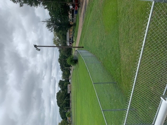 A lush grass field with an L-shaped section of chain-link fencing in the foreground and center, a road further back towards the right, and trees in the background under a cloudy sky.
