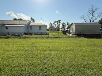 An open grass field with a chain-link fence in the center, houses beyond the fence, and trees further in the distance under a clear blue sky.