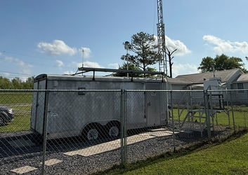 A metal camper trailer in the center of a fenced-in gravel plot within a grass field; trees, houses, and clouds are in the background.