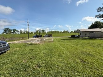 A vast grass field with a house to the right, a car and pavement to the left, and a grassy river levee in the background beyond the road.