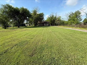 A vast grass field with a gravel driveway to the right, residential home in the background, and trees bordering the background.