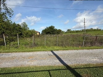 A gravel driveway in the foreground, flanked by grass on either side, with a fence on the far side of the driveway separating it from a field of tall vegetation under a cloudy blue sky.
