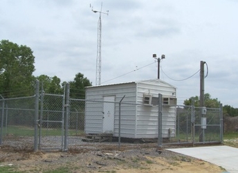 A small white shed inside a chain-link fenced-in area with grass and trees in the background, along with dirt and a sidewalk in the foreground under a cloudy sky.