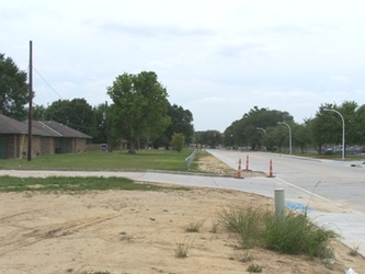 A patch of dirt in the foreground and grass in the background, divided by a horizontal road that joins a vertical road on the right side; there are trees in the background and a house to the left.