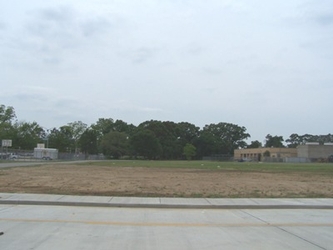 A road in the foreground, bordered by a sidewalk and then a mostly bare dirt/grass field beyond that with trees and buildings in the background under a cloudy sky.