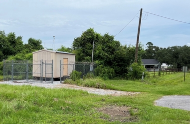 A grass field with a gravel and pavement driveway leading to a chain-link fenced-in area containing a small tan shed with trees and homes in the background.