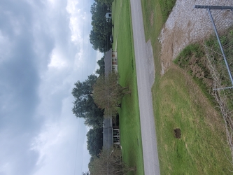  A residential area with homes, trees, and yards in the background, a road running horizontally down the center, and a grass area in the foreground with a gravel driveway in the middle.