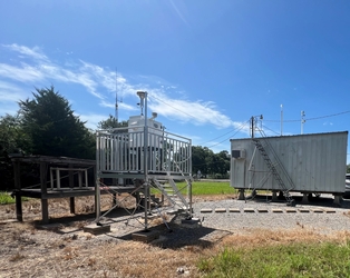 A small building on the right and a metal platform with air sampling equipment on the left; grass, gravel, and stepping stones are in the foreground and trees are in the background.