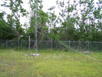 A grassy area with a chain-link fence running horizontally down the center, a narrow metal tower towards the foreground, and trees in the background.