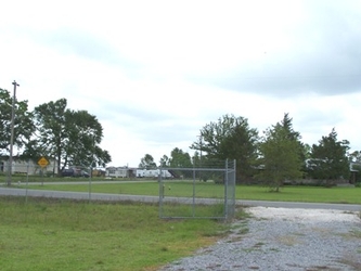 Grass in the foreground with a gravel driveway to the right, a road and chain-link fence running horizontally down the center, and more grass in the background along with trees and homes further back.