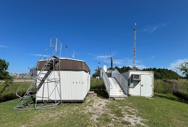 A small building on the left with a staircase leading to the roof, a smaller building on the right next to a metal platform with air sampling equipment, and a clear blue sky overhead.