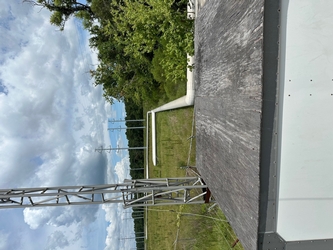The backside and roof of a small building with a metal meteorological tower on the left, grass in the background, and trees in the far distance under a cloudy sky.