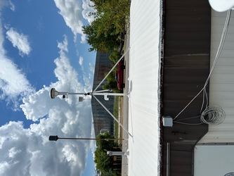 The side and roof of a small building with air sampling equipment on the roof, a large structure in the far distance, and a road with cars and trees in the center under a cloudy blue sky.