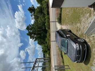 A gray minivan in the foreground on a gravel driveway within a grassy area, a chain-link fence separating the grass area from a road, and trees in the background under a cloudy blue sky.