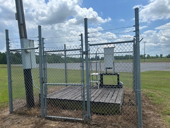 A fenced in area holding a wooden platform with a piece of air monitoring equipment, with a parking lot and trees in the background.
