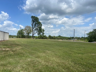 A far-reaching grassy field with a shed on the far left and trees lining in the horizon under a cloudy blue sky.