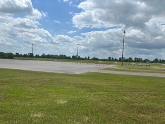 An empty, large concrete parking lot with grass surrounding all sides; there are electrical poles beyond the lot and trees lining the horizon under a partly cloudy sky.
