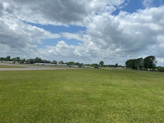 A large grassy field with buildings and cars inside a parking lot on the distant left, a concrete drive along the lefthand side, and trees throughout the background under a cloudy blue sky.