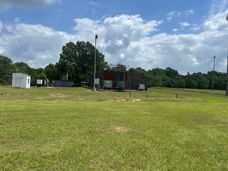 A grassy field with a large, black, metal industrial structure and trees in the background.